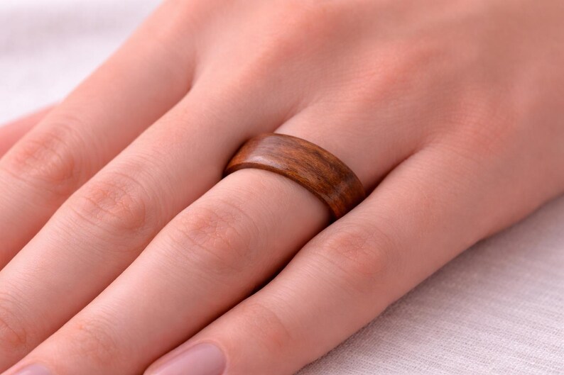 May include: Close-up of a wooden ring with a natural wood grain pattern. The ring is a warm brown color and is worn on a finger. The ring is a simple band, showcasing the wood's natural beauty.