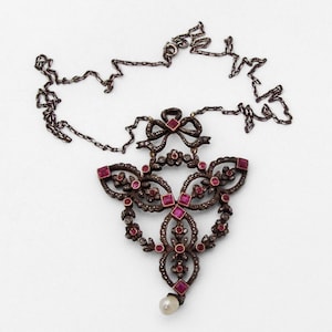 Op de afbeelding: Een zilveren ketting met een hanger met een delicaat, ingewikkeld ontwerp van verweven bloemmotieven en robijnrode edelstenen. De hanger is versierd met een strik en een enkele parel hangt aan de onderkant.