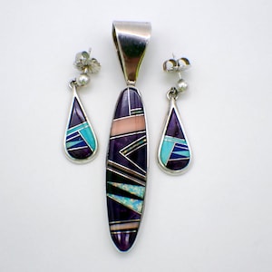 May include: A silver pendant and earring set with a geometric design. The pendant is long and oval, with a silver bail. The earrings and pendant feature purple, turquoise, pink, and white inlaid stones. The set is displayed on a white background.