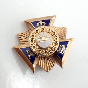 May include: A gold-coloured pin with a central circular design featuring a sunrise over a white horizon. The pin has blue enamel accents and the Greek letters Alpha and Omega. The pin has a crown at the top.