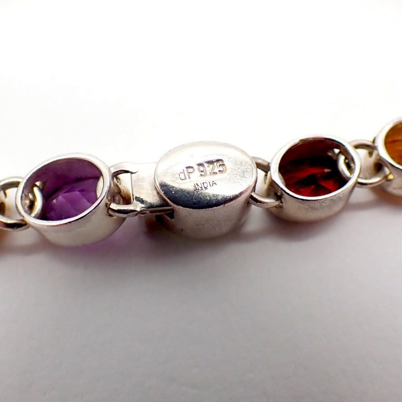 Puede incluir: Un brazalete de plata con un cierre marcado con "dp 925 INDIA". El brazalete presenta piedras preciosas redondas y coloridas en tonos de morado, rojo y amarillo.