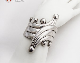 Puede incluir: Un brazalete de plata con un diseño ondulado y acentos redondeados. El brazalete está hecho de múltiples capas de plata y tiene un estilo único y moderno.