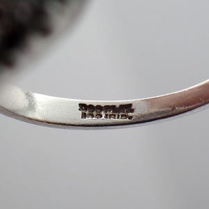 May include: Close-up of a silver ring with a hallmark that reads "925 ITALY". The ring's band is smooth and polished, reflecting the light. The ring is likely a piece of jewelry.