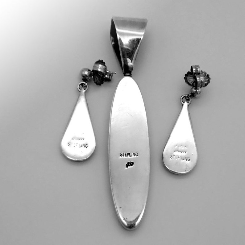 May include: A silver pendant and matching earrings set. The pendant is elongated with a curved top, and the earrings are teardrop-shaped. Each piece is stamped with "STERLING". The set is displayed on a light gray surface.