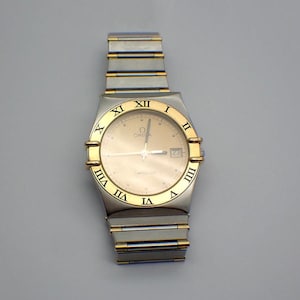 May include: A gold and silver two-tone Omega Constellation wristwatch with a white face and Roman numeral hour markers. The watch has a date window at the 3 o'clock position.