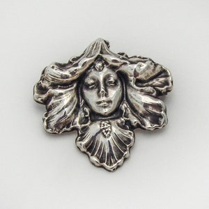 May include: A silver brooch featuring a woman's face with flowing hair, set within a stylized floral design.