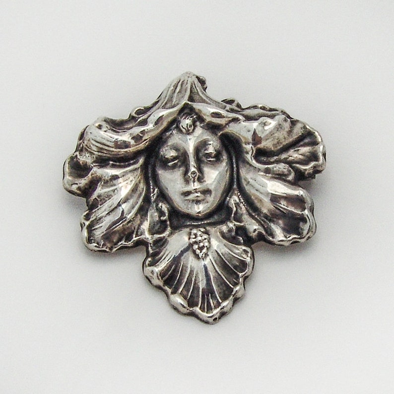 May include: A silver brooch featuring a woman's face with flowing hair, set within a stylized floral design.