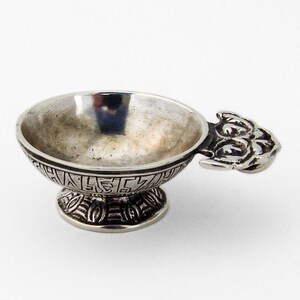 Op de afbeelding: Een zilveren kom met een handvat in de vorm van een gestileerd gezicht. De kom heeft een decoratief patroon rond de rand en de basis.