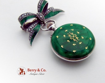 May include: A green enamel pocket watch with a gold floral design and a silver bow brooch with pearl accents.