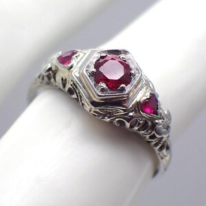 Peut inclure: Une bague en argent ornée de trois pierres rouges. La pierre centrale est un grand rubis facetté serti dans une lunette hexagonale. Deux petits rubis en forme de cœur flanquent la pierre centrale. La monture présente des motifs floraux et volutes.