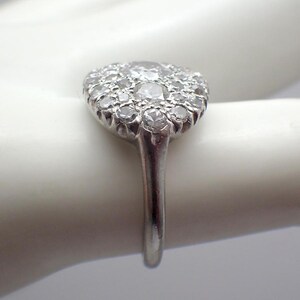 May include: A silver ring featuring a cluster of small, round diamonds set in a circular pattern. The ring's band is a simple, polished silver. The diamonds sparkle, catching the light.