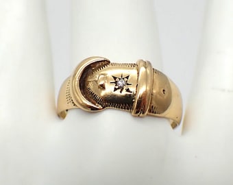 May include: A gold ring featuring a buckle design with a small diamond set in the centre. The ring is displayed against a white background.