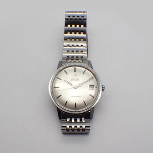 May include: A silver Omega Seamaster automatic wristwatch with a silver dial and date display. The watch has a stainless steel band with a grid-like design. The watch is displayed against a light grey background.