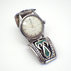 May include: Vintage silver-toned wristwatch with a white face and the word "Earlbert" printed on the dial. The watch features a decorative band with turquoise-colored inlays and an ornate design. The watch is an automatic 21 jewels.