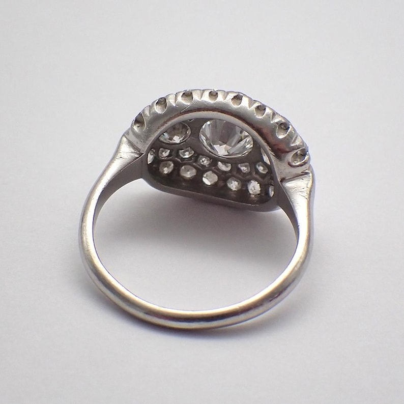 May include: A silver ring with a large, central, round-cut diamond surrounded by smaller diamonds. The ring's band is smooth and rounded, and the setting features a detailed, scalloped edge. The ring is set against a white background.