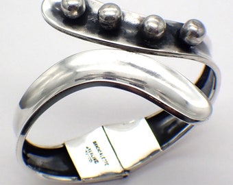 Puede incluir: Un brazalete de plata con un diseño curvo y cuatro acentos redondos. El brazalete tiene un cierre con bisagra y está marcado con "Birkallerst Sterling 925".