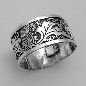 Op de afbeelding: Een zilveren armband met een ingewikkeld bloemen- en paisley-ontwerp. De armband is gemaakt van metaal en heeft een scharniergesp.