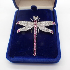 May include: A dragonfly brooch crafted from silver-toned metal, featuring a long body set with a vertical row of oval-cut pink rubies. The four wings are encrusted with numerous small clear white stones and accented with pink rubies at the tips.
