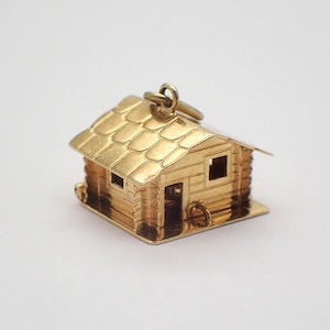 May include: A gold-colored charm in the shape of a log cabin. The charm has a detailed roof, windows, and a door.