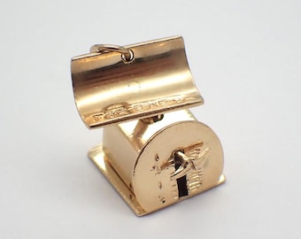 Op de afbeelding: Gouden bedel in de vorm van een vintage weegschaal. De weegschaal heeft een ronde basis en een platte bovenkant met een gebogen overhang. De bedel heeft een lus om aan een armband of ketting te bevestigen.