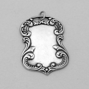 May include: A silver pendant with an ornate, scrolled design. The pendant is blank and has a loop at the top for attaching a chain.