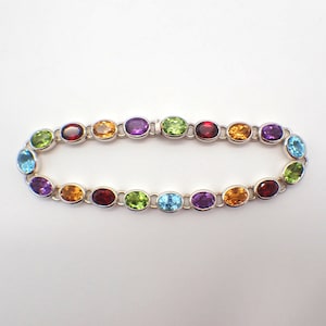 Puede incluir: Un brazalete de plata con piedras preciosas ovaladas de varios colores, incluyendo rojo, naranja, amarillo, verde, azul y morado.
