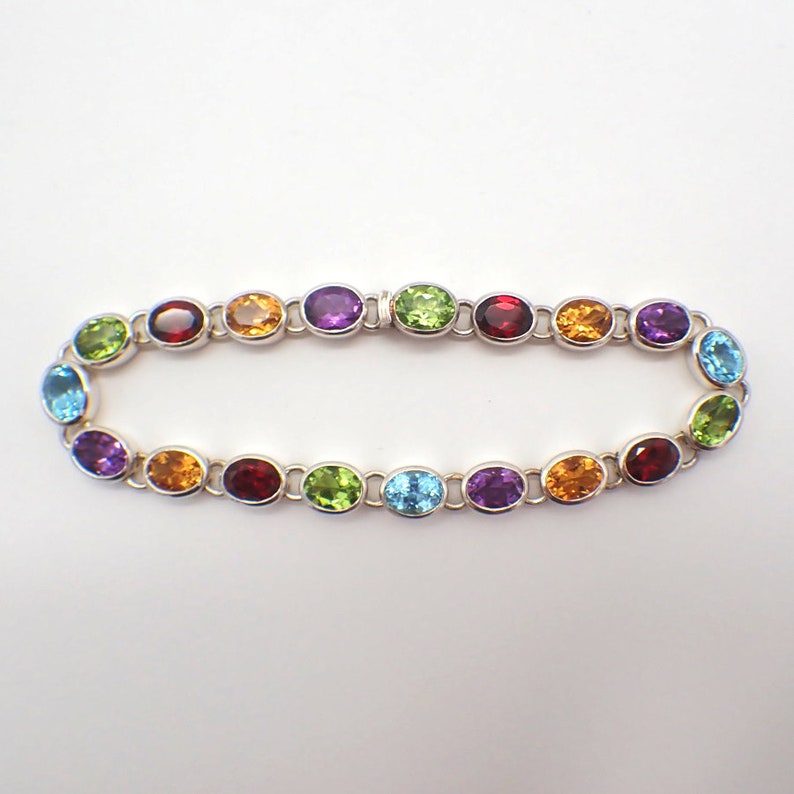 Puede incluir: Un brazalete de plata con piedras preciosas ovaladas de varios colores, incluyendo rojo, naranja, amarillo, verde, azul y morado.