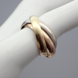 May include: A three-band ring with gold, rose gold, and silver bands. The bands are intertwined, creating a unique and elegant design. The ring is shown on a white surface.