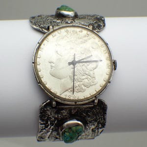 May include: A silver wristwatch with a coin face, featuring a detailed profile. The watch has a textured silver band with turquoise accents. The watch face displays the year 1884. The watch is a unique piece of jewelry.