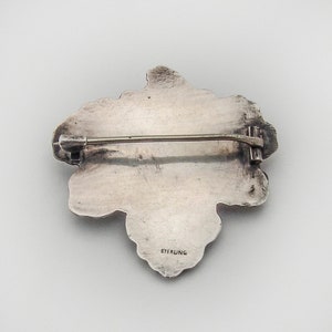 May include: A silver brooch in the shape of a cluster of grapes. The back of the brooch has a pin and a clasp, and the words "STERLING" are stamped on the back.