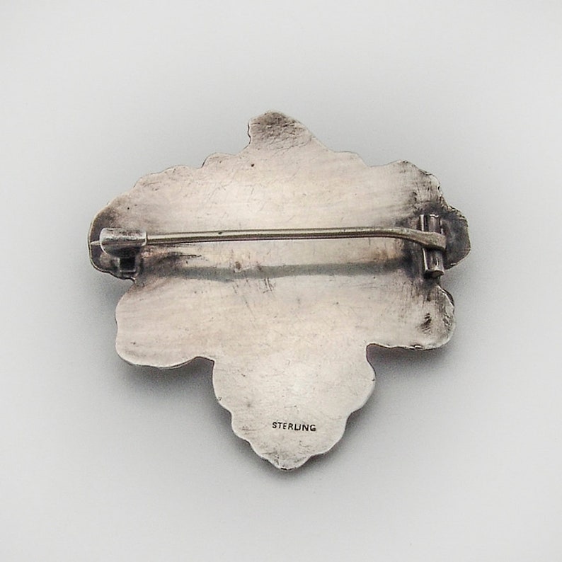 May include: A silver brooch in the shape of a cluster of grapes. The back of the brooch has a pin and a clasp, and the words "STERLING" are stamped on the back.