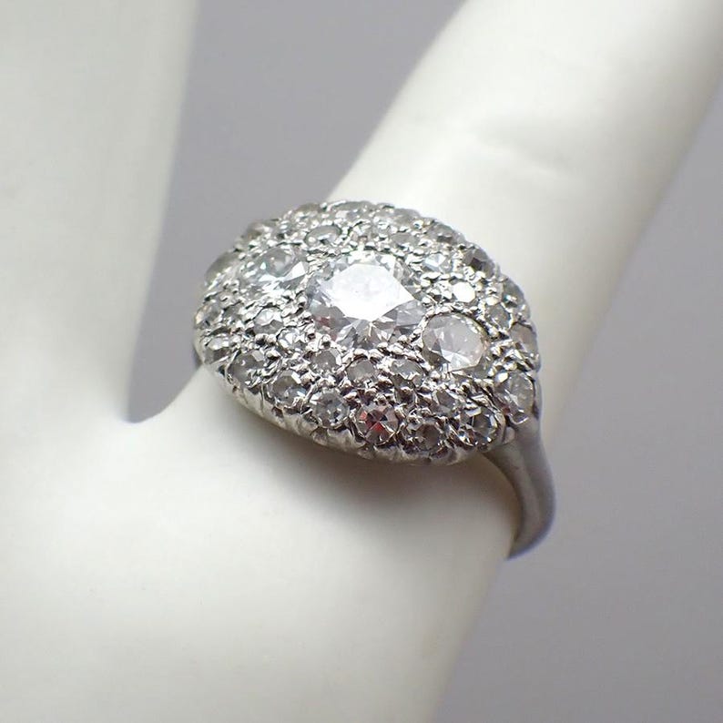 May include: A silver ring featuring a cluster of small, clear gemstones surrounding a larger, central stone. The ring is set on a white background, showcasing the intricate details of the jewelry.