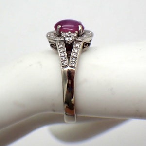 May include: A silver ring with a pink oval gemstone surrounded by smaller clear gemstones. The ring has a delicate, intricate design.