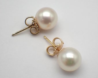 Puede incluir: Un par de pendientes de botón con perlas redondas y brillantes de color blanquecino. Las perlas están engarzadas en postes de oro con detalles curvos decorativos. Los pendientes se muestran sobre una superficie blanca.