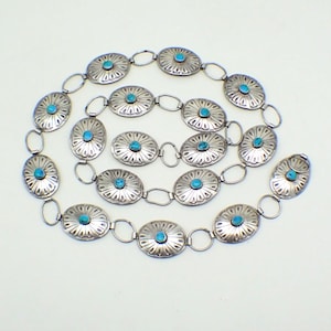 May include: A silver-coloured belt with oval links, each with a turquoise-coloured stone centre. The links are joined by small, circular rings. The belt's design incorporates a floral pattern around each stone, creating a decorative accessory.