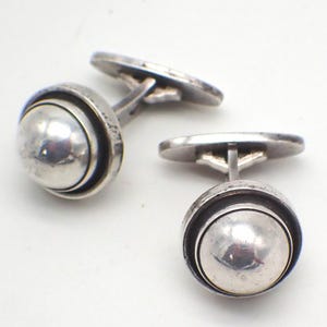 Può includere: Un paio di gemelli in argento. Ogni gemello presenta una sfera argentata lucida e arrotondata, incorniciata in un cerchio. I gemelli hanno un design classico con chiusura a T. Il metallo ha un aspetto leggermente invecchiato.