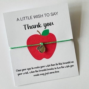 Könnte beinhalten: Eine weiße Karte mit dem Text "A Little Wish To Say Thank You" und einem roten Apfel mit einem grünen Blatt und einem silbernen Apfel-Anhänger, der an einem grünen Schnur-Armband befestigt ist. Der Text auf der Karte lautet: "Schließe deine Augen, um dir etwas zu wünschen, und binde dann dieses Armband an dein Handgelenk. Wenn das Armband in zwei Teile zerbricht, kann der Wunsch, den du dir gemacht hast, in Erfüllung gehen."