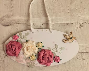 May include: Oval-shaped white wooden wall hanging decorated with paper flowers in shades of pink, cream, and yellow. A small wooden bird is attached to the right side. A white ribbon is attached for hanging.