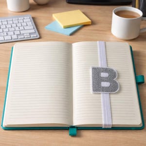 May include: An open teal notebook with lined pages, a silver glitter letter "B" on a white elastic band, and a teal ribbon bookmark. The notebook is on a wooden desk with office supplies.