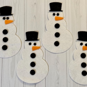 May include: Four white felt snowman decorations with black top hats, black button eyes and bodies, and orange carrot noses. The snowmen are arranged on a light wood-grain background.