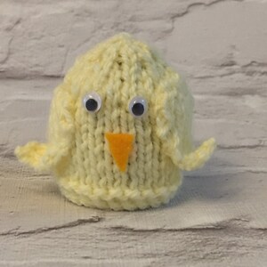 May include: A yellow knitted chick with white googly eyes and an orange felt beak. The chick is sitting on a white textured surface.