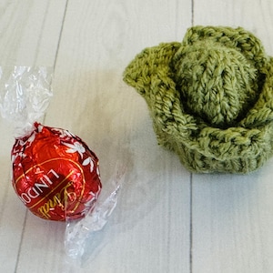 May include: A red foil-wrapped Lindt chocolate truffle and a handmade green knitted cabbage. The truffle has white floral accents and the word "Lindt" printed on it. The cabbage is made of green yarn.