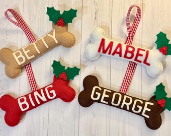 May include: Four felt dog bone ornaments with holly and berry accents. The bones are decorated with the names "Betty", "Mabel", "Bing", and "George".