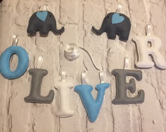 Könnte beinhalten: Ein Filzbanner mit dem Namen "OLIVE", der in blauen und grauen Buchstaben ausgeschrieben ist. Zwei graue Filzelefanten mit blauen Herzen befinden sich auf beiden Seiten des Banners.