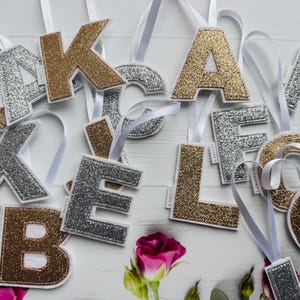 May include: Glitter-covered fabric letters in gold, silver, and brown, each with a white ribbon for hanging. The letters are arranged on a white surface with a pink rose and greenery, perfect for personalized decor.