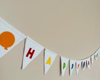 Op de afbeelding: Witte vlag van vilt met de woorden "Happy Birthday" in regenboogkleuren. Elke letter staat op een aparte driehoekige vlag. Op de eerste vlag staat een oranje ballon.
