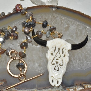 May include: A gold chain necklace with a carved white bone bull skull pendant with black horns. The necklace is made with faceted gray and brown gemstones.