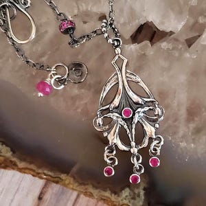 Puede incluir: Un collar de plata con un colgante con un diseño floral y piedras preciosas rosas. El colgante tiene un diseño delicado e intrincado con múltiples capas de plata.