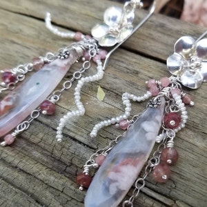 May include: A pair of silver dangle earrings featuring a flower charm and a long, teardrop-shaped agate stone. The earrings are decorated with small pink and white beads, and silver chains.