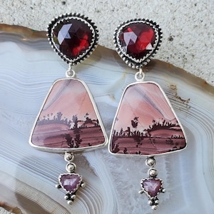 May include: A pair of dangle earrings featuring faceted deep red garnet stones at the top, set in silver. Below are trapezoidal landscape jasper stones with brown and pink bands, finished with small, faceted, triangular pinkish-red stones at the base.